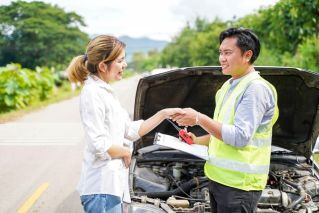 A man and a woman shaking hands in front of a broken down car. He is wearing a safety vest