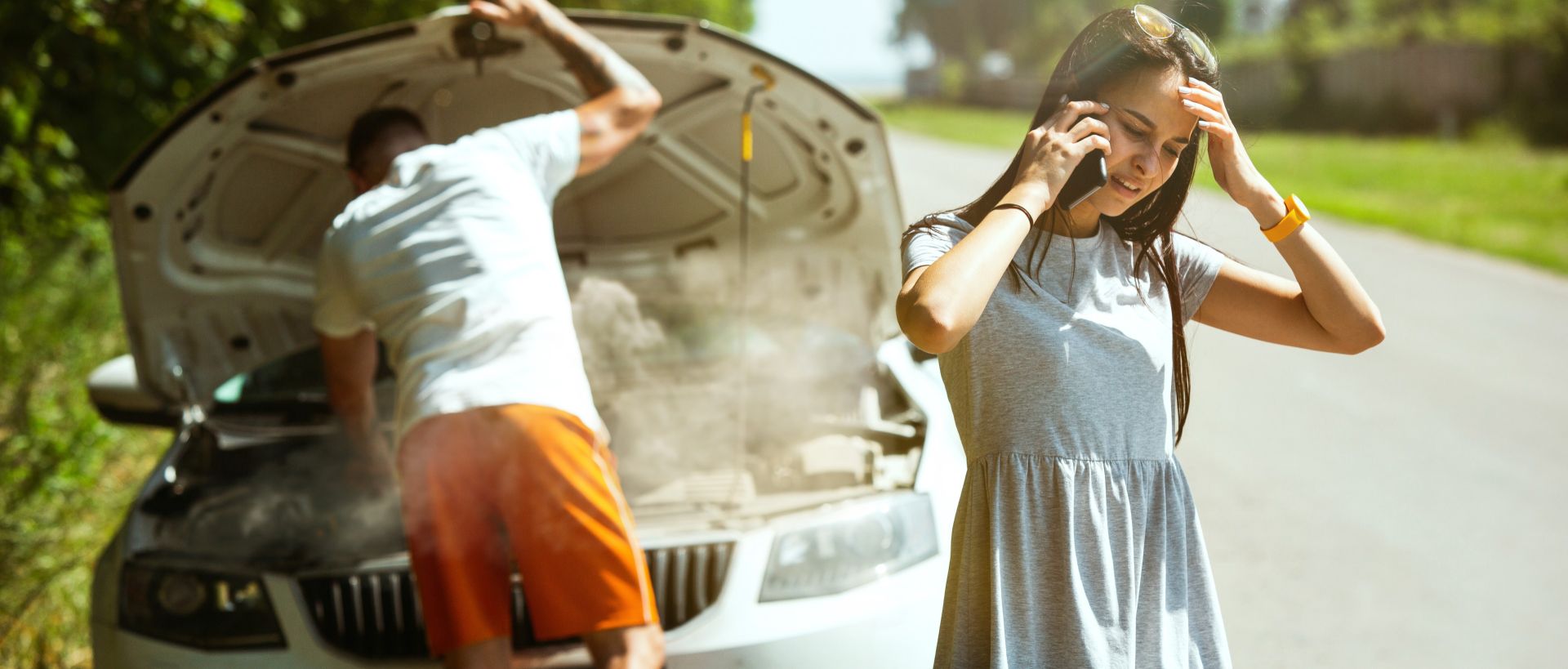 A couple in front of broken down car; he is looking at the engine and she is on her cellphone, looking stressed